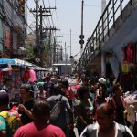 Mercado de rua movimentado, com pessoas circulando entre bancas coloridas.