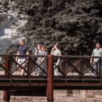 Adultos caminham por uma ponte de madeira em meio à natureza durante um passeio.