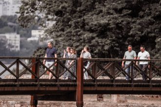 Adultos caminham por uma ponte de madeira em meio à natureza durante um passeio.